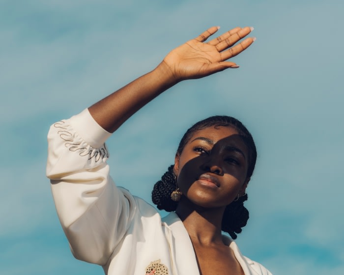 Portrait of a woman with braided hair shielding her face from sunlight against a blue sky, wearing a white jacket, and gold earrings.