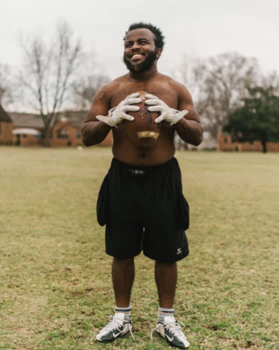 Adult male athlete in sports uniform and gloves holding football on an athletic field outdoors on an overcast day.