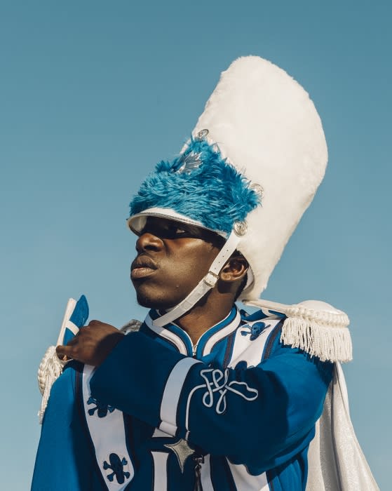 Portrait of a black adult male in a blue and white marching band uniform with a tall hat against a clear blue sky.