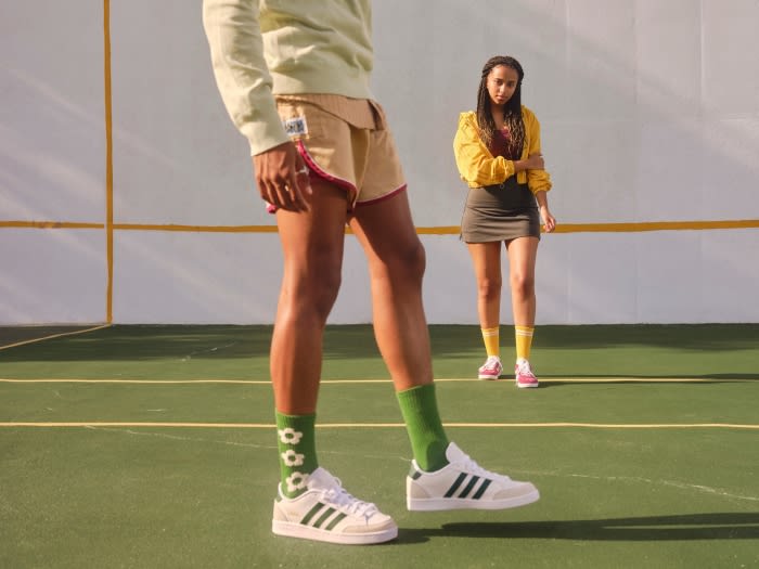Two people in sports clothing outdoors on a tennis court. One person is standing with legs slightly raised, wearing white sneakers.