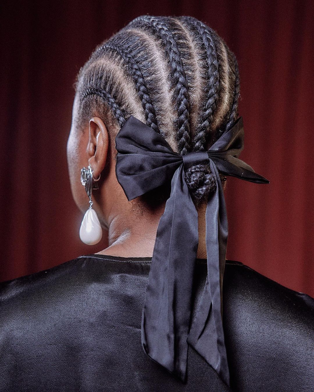 Adult woman with braided hair pulled up into a bun tied with a black ribbon, facing away from the camera, wearing dangling earrings.