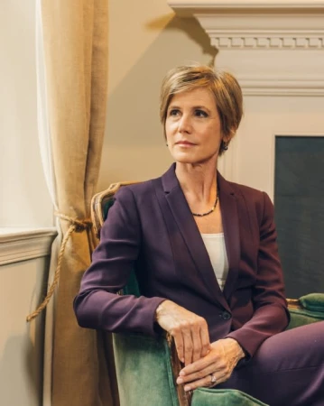 Indoor portrait of a stylish adult woman in a purple business suit sitting in a green armchair, looking away from the camera.
