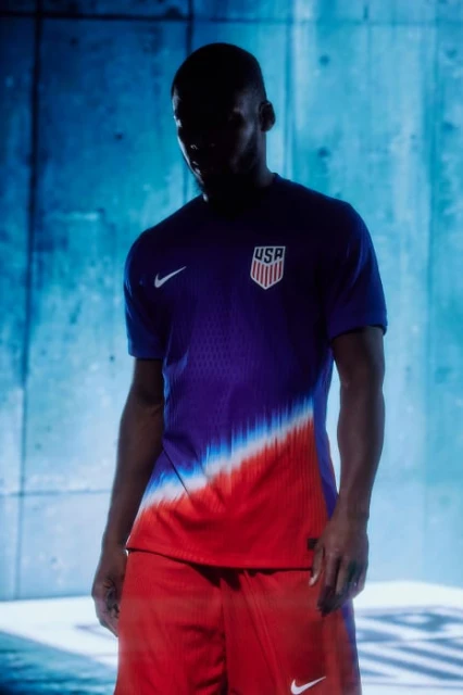 A person wearing a USA soccer jersey and shorts stands in front of a textured blue wall with dark lighting and the Nike logo visible.