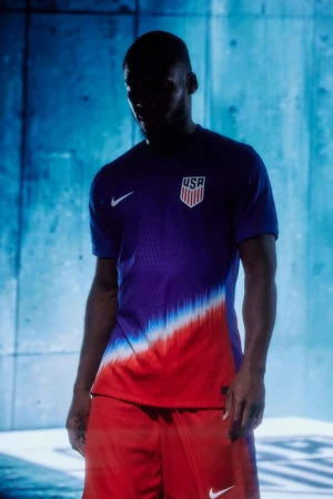 A person wearing a USA soccer jersey and shorts stands in front of a textured blue wall with dark lighting and the Nike logo visible.