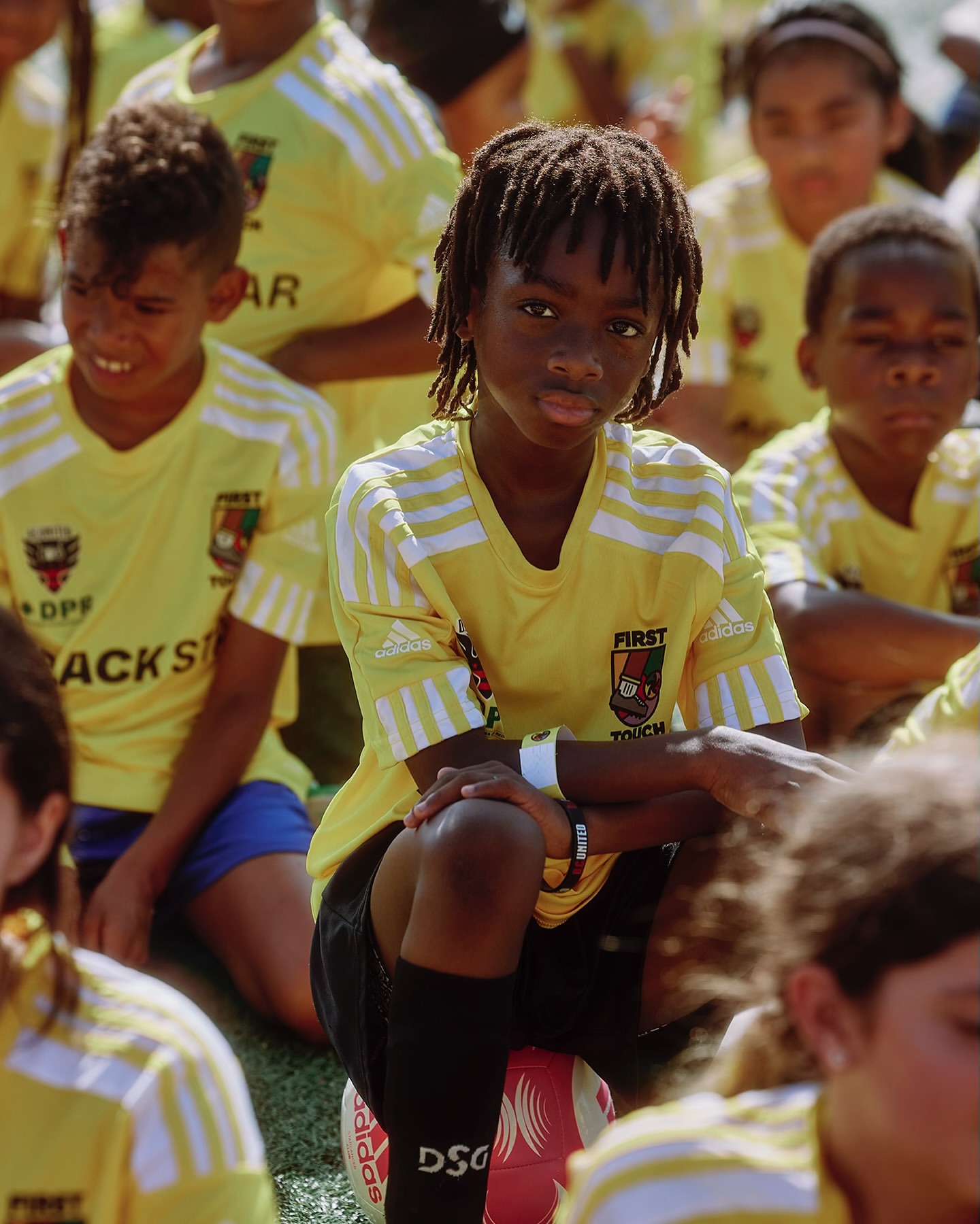 Youth soccer team members in yellow and white uniforms gather on the field, featuring a young Black boy seated on a soccer ball.