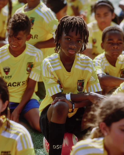 Youth soccer team members in yellow and white uniforms gather on the field, featuring a young Black boy seated on a soccer ball.