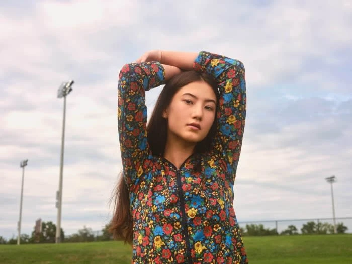 Portrait of an Asian young woman in a floral jacket, posing outdoors in front of a grassy field and a cloudy sky, creating a natural feel.