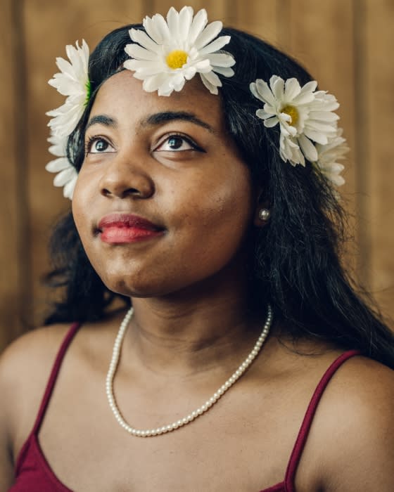 Portrait of a woman with dark hair and a daisy flower crown, wearing a maroon tank top and pearl necklace, gazing upwards.