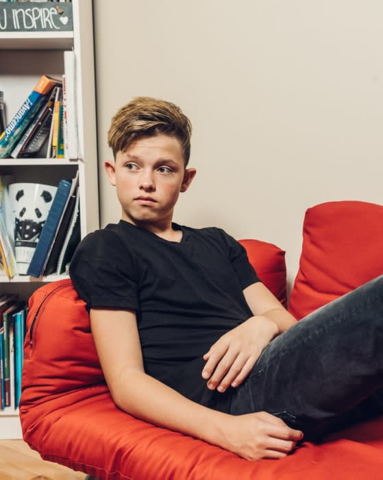 Teenage male in a black t-shirt and jeans lounges on a red beanbag next to a bookshelf in an indoor setting with soft lighting.
