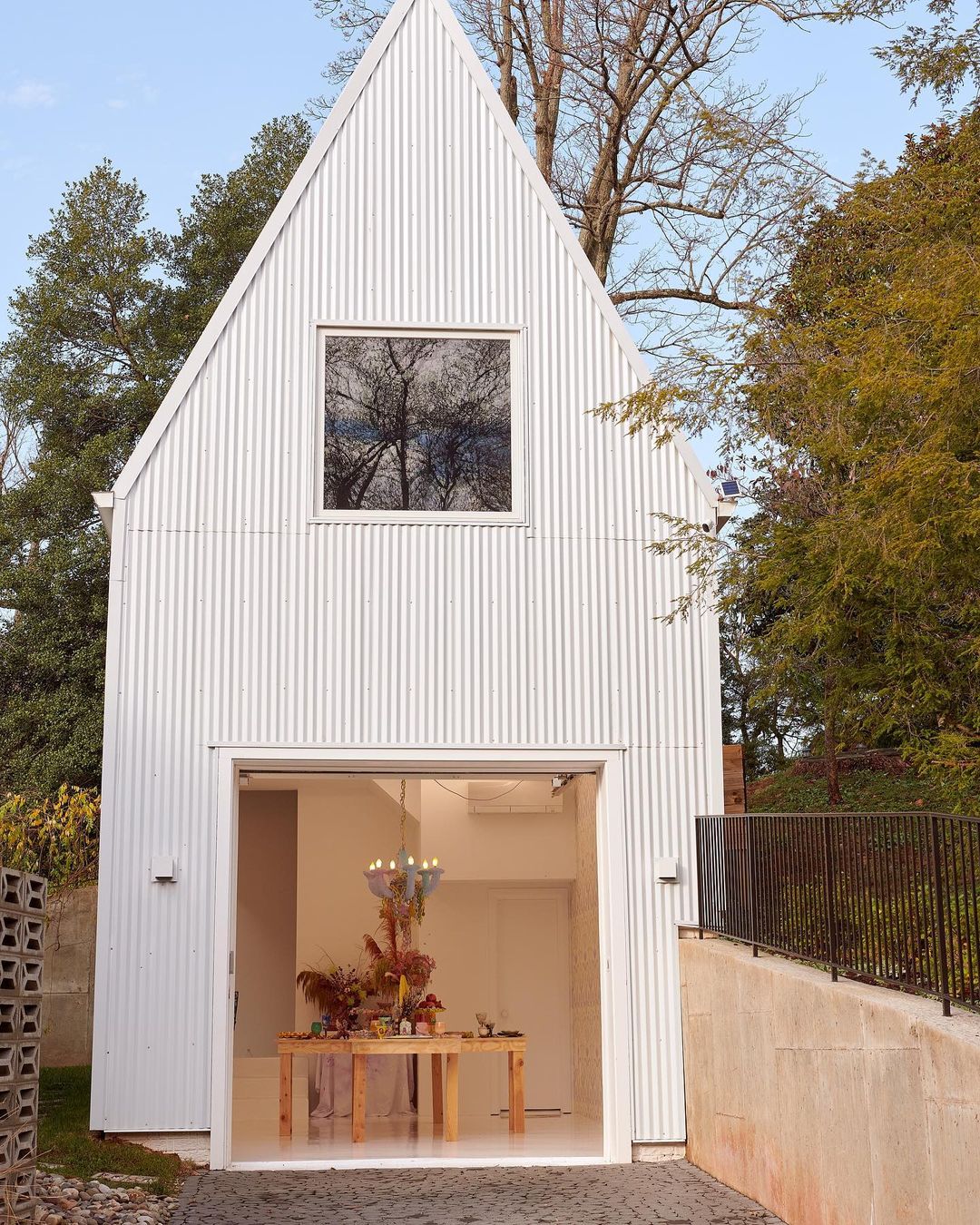 Exterior shot of a modern white barn-style building with a window and glass door, displaying a table and floral arrangement indoors.