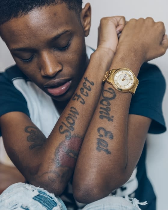 Close-up of a young adult with tattoos on their forearms wearing a gold watch, set against a neutral background in an indoor environment.
