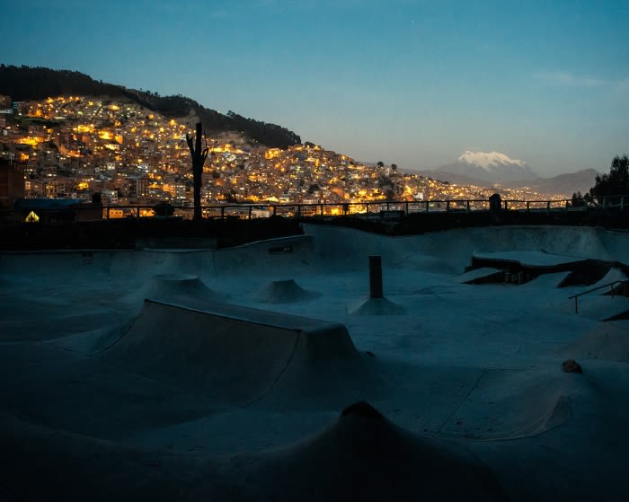 Nighttime view of an empty skatepark bowl with a distant city on a hill illuminated in warm lights, and a snow-capped mountain visible in background.