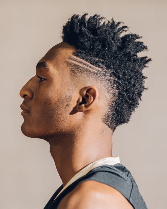Profile portrait of a Black male with a shaved head and a textured mohawk with parallel shaved lines, against a beige backdrop.