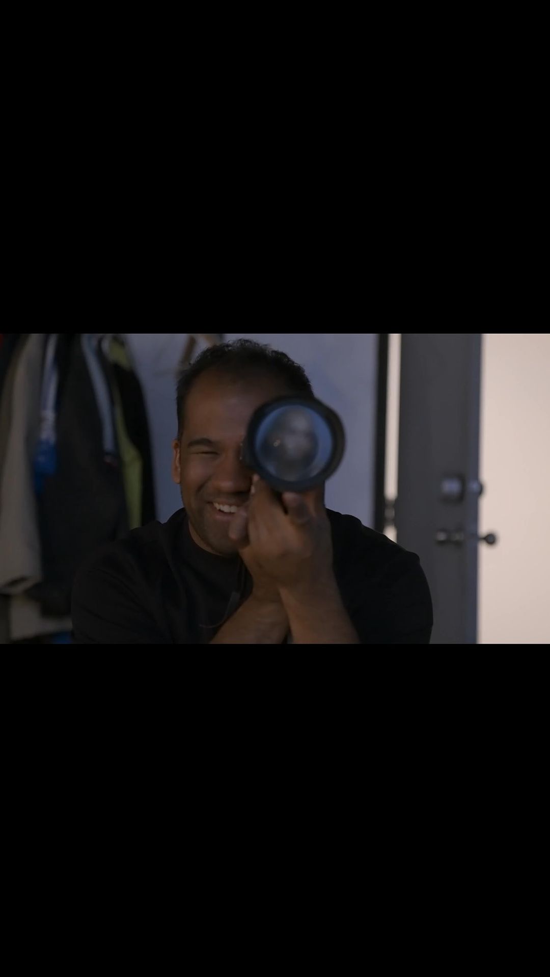 Smiling African-American man holds a black object to his eye in an indoor setting with a partially visible white door.