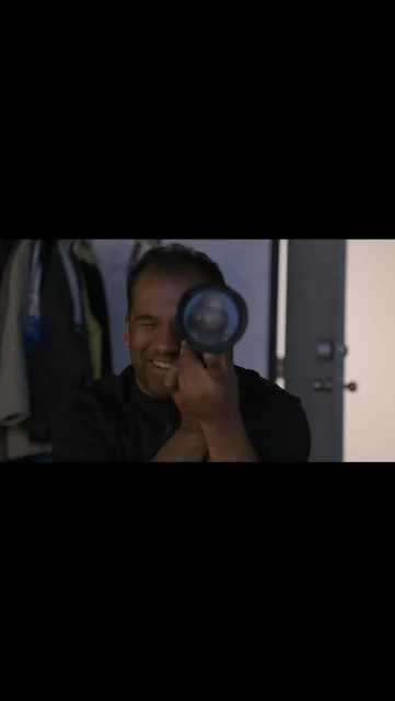 Smiling African-American man holds a black object to his eye in an indoor setting with a partially visible white door.
