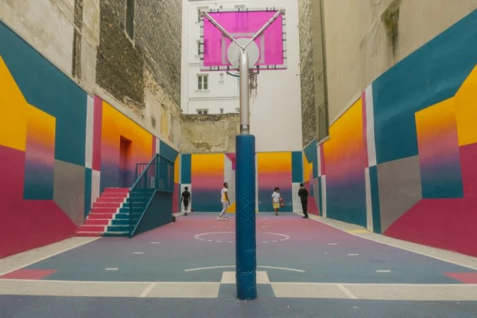 Outdoor basketball court surrounded by mural-covered walls; kids play on the blue and pink painted surface with pink basketball hoop.