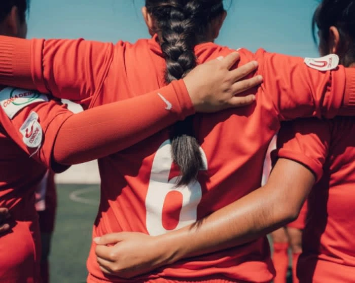 Three female soccer players huddle with arms around each other, wearing red jerseys on a sunny day. The focus is on team unity.
