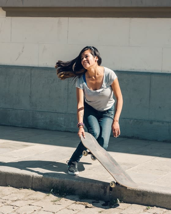 Smiling woman holding a skateboard on city street. Casual outfit, dark hair, urban daytime setting, good natural light, neutral tones.