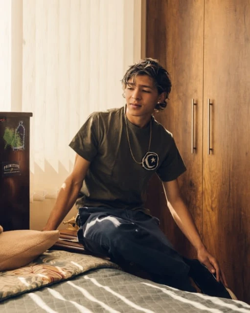 Young adult male with silver hair, wearing a green t-shirt and jeans, sits on a bed with a brown wood wardrobe in the background.