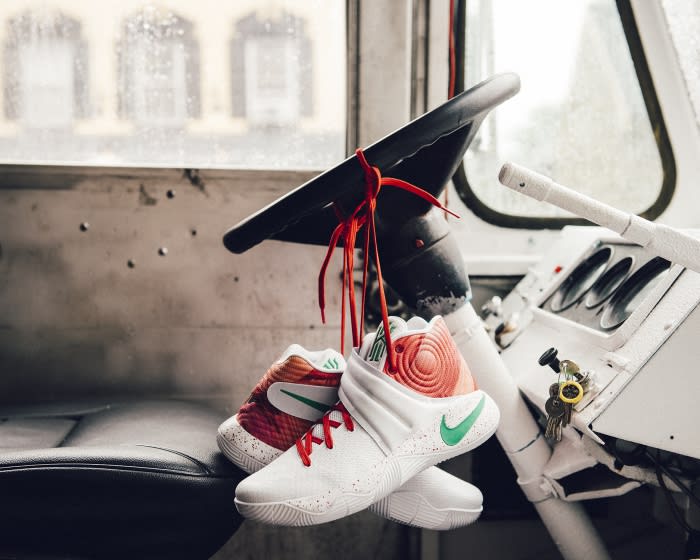 White, orange, and green athletic shoes tied to a black steering wheel inside of vehicle with view through rain-covered windshield.