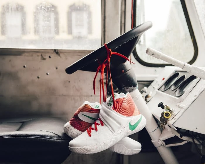 White, orange, and green athletic shoes tied to a black steering wheel inside of vehicle with view through rain-covered windshield.