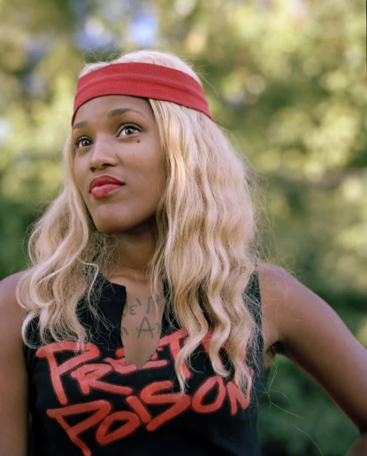 Portrait of a Black woman with blond wavy hair, red headband, and a black tank top in an outdoor setting with blurred foliage.