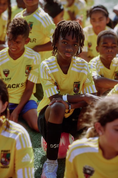 A group of kids in yellow soccer jerseys, one boy with dreadlocks front and center, leans on a soccer ball at eye level in the daytime.