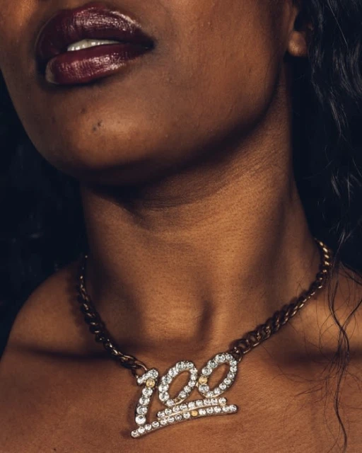 Close-up of a dark-skinned woman with dark lipstick wearing a gold chain necklace with a '100' pendant. The focus is on beauty and jewelry.