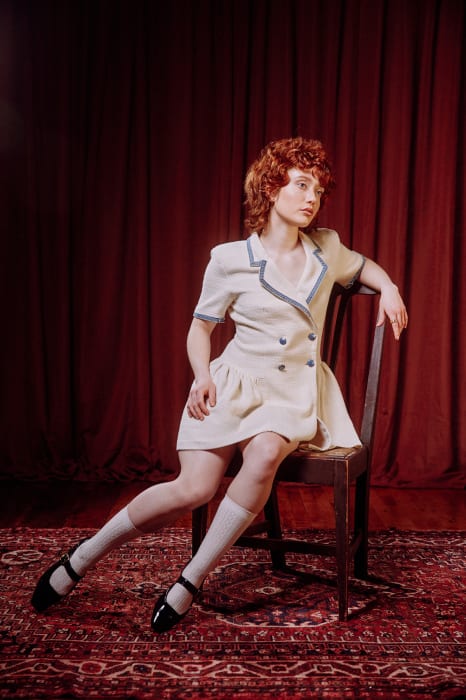 A fair-skinned young woman with auburn hair sits in a white dress and Mary Jane shoes in front of a red curtain and patterned rug.