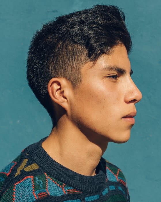 Profile shot of a young adult male with dark hair wearing a patterned sweater against a solid teal background in natural lighting.