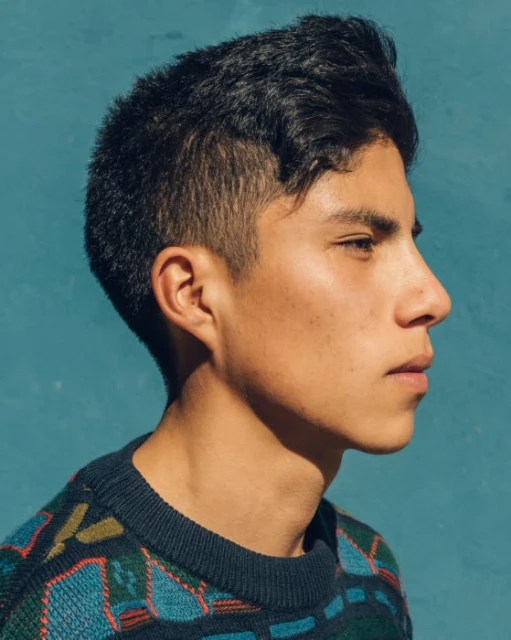 Profile shot of a young adult male with dark hair wearing a patterned sweater against a solid teal background in natural lighting.