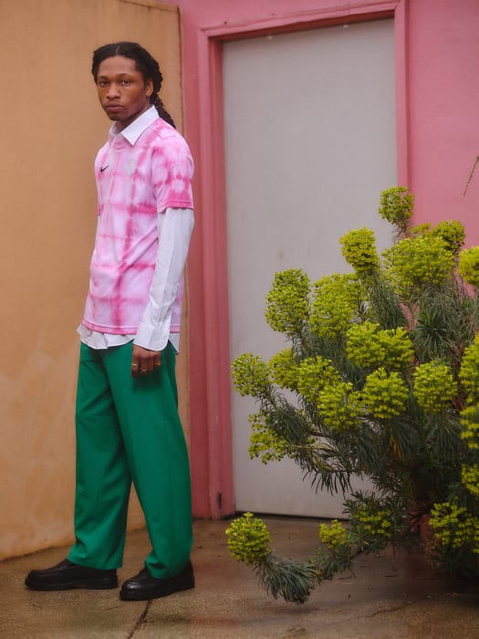 A young adult poses next to a white door and bright green plant wearing a tie-dye shirt and green pants in an outdoor setting.