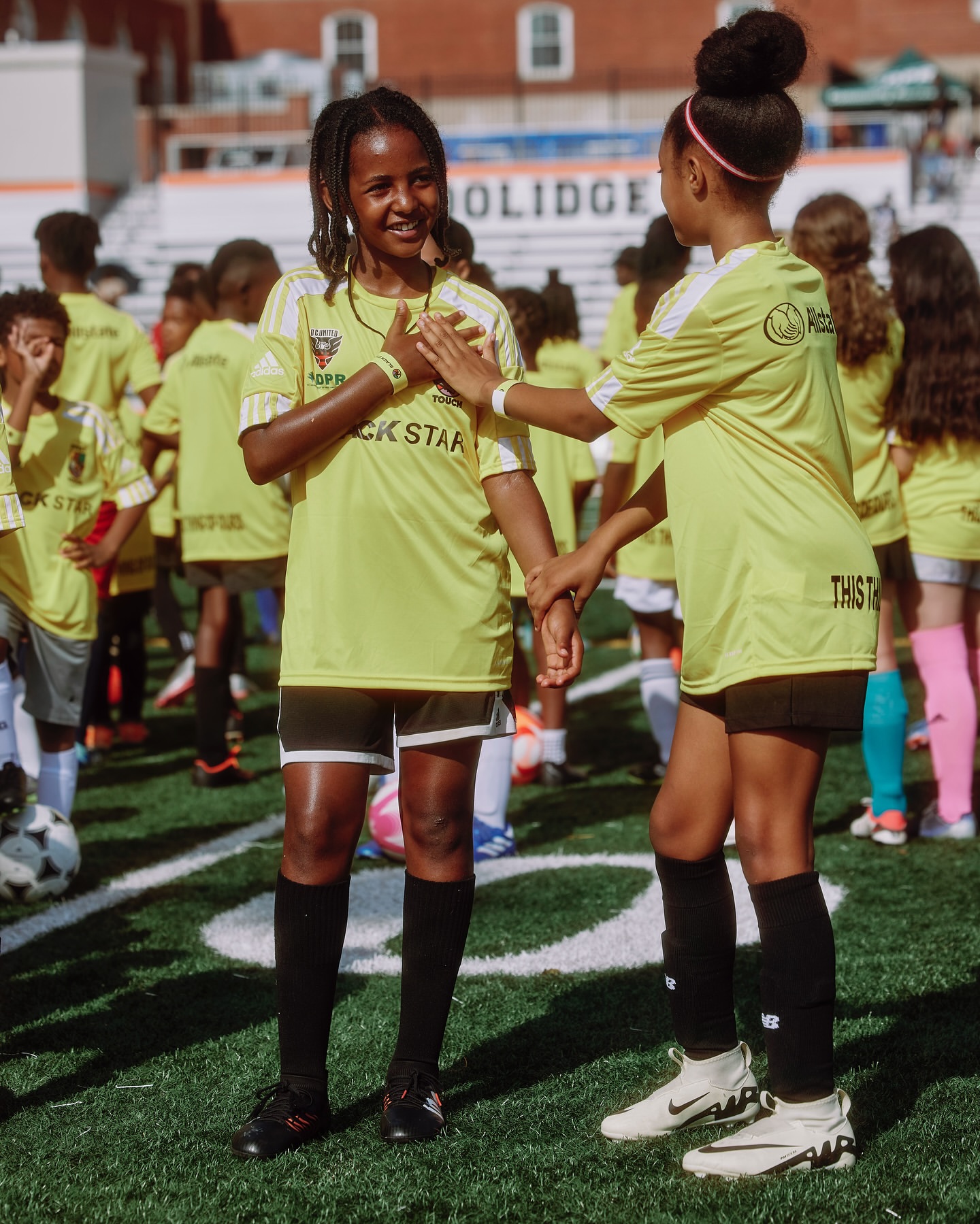 Youth soccer team gathers on the field, two girls in yellow jerseys stand close, with other children and stadium seating in the background.