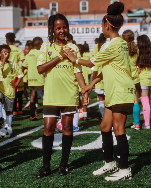 Youth soccer team gathers on the field, two girls in yellow jerseys stand close, with other children and stadium seating in the background.