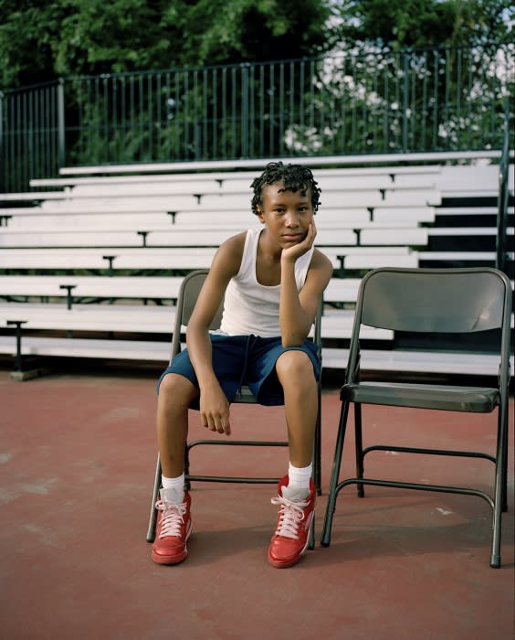Young boy sits on a folding chair, wearing red sneakers, outdoors near bleachers and a green tree line in a medium shot.