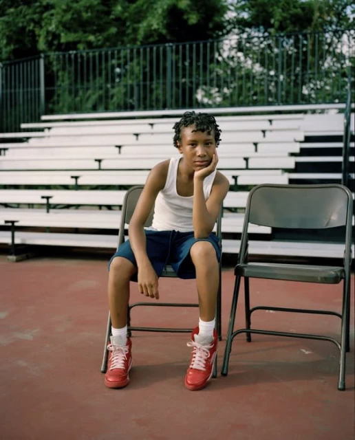 Young boy sits on a folding chair, wearing red sneakers, outdoors near bleachers and a green tree line in a medium shot.