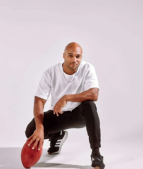 Athletic, bald man with an American football crouches in a photo studio. The man wears a white t-shirt, black pants, and black tennis shoes.