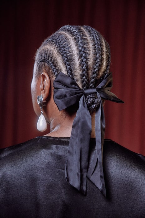 Rear view portrait of a Black woman with braided hair and a black satin bow, accessorized with silver and pearl drop earrings, against a maroon curtain.