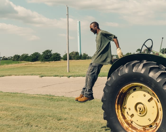 Man leaning on vintage tractor in grassy field on a sunny day. Tractor has green and yellow paint with black tires in a rural scene.