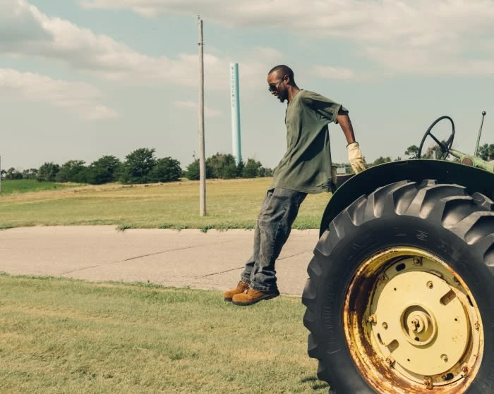 Man leaning on vintage tractor in grassy field on a sunny day. Tractor has green and yellow paint with black tires in a rural scene.