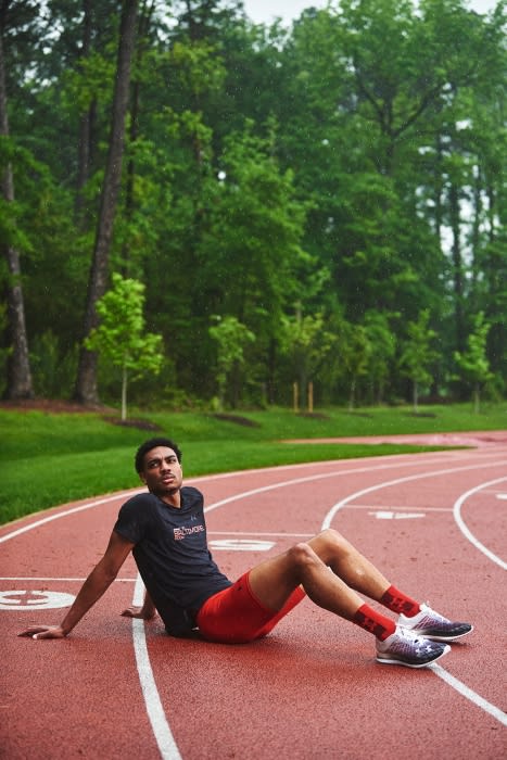 Adult male athlete rests on a red running track, wearing black shirt, red shorts, socks and running shoes, with green trees behind.