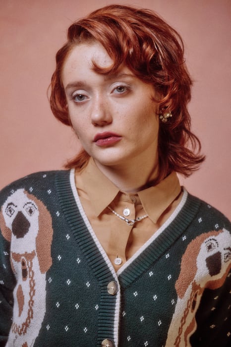 Portrait of a red-haired woman in a dog-patterned cardigan and tan collared shirt against a pink background in soft, studio lighting.