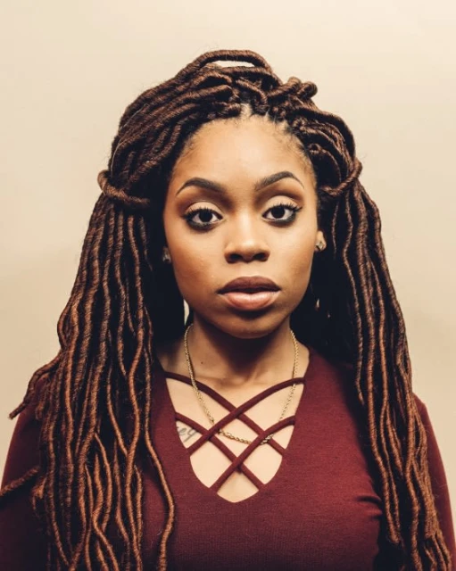 Portrait of a woman with long brown faux locs, wearing a maroon top and gold chain necklace, against a neutral background.