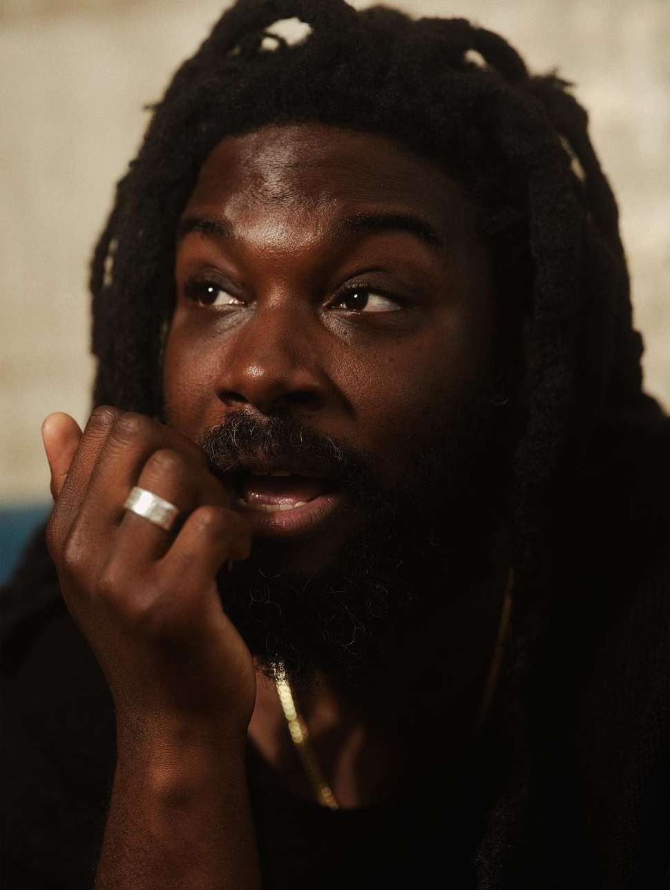 Portrait of a Black man with dreadlocks, wearing a black shirt and gold chain, with a ring on his finger, indoors with neutral walls.