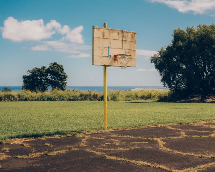Outdoor basketball court features a weathered backboard, yellow pole, and net with green grass and blue sky with clouds in the background.