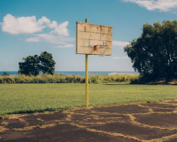 Outdoor basketball court features a weathered backboard, yellow pole, and net with green grass and blue sky with clouds in the background.