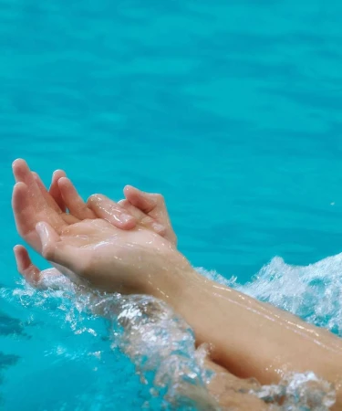 Close-up shot of two hands gently submerged in turquoise water, creating splashes and ripples around the wrists and fingers.