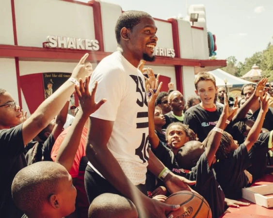Kevin Durant smiles among a crowd of children outdoors, wearing a white t-shirt and holding a basketball, with an amusement park background.