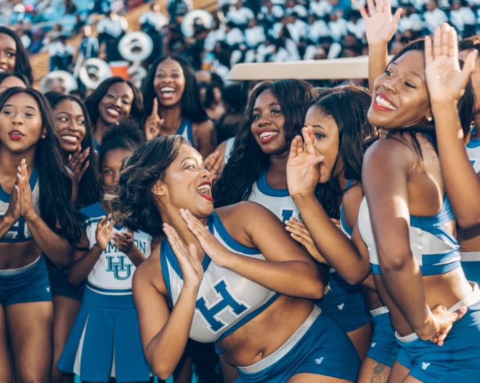 Enthusiastic group of diverse female cheerleaders in blue and white uniforms perform at an outdoor daytime sporting event with stadium seating.