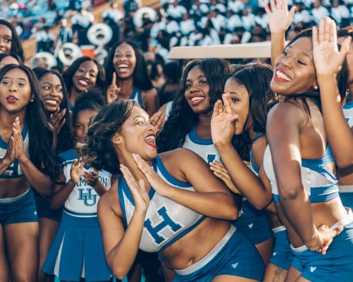 Enthusiastic group of diverse female cheerleaders in blue and white uniforms perform at an outdoor daytime sporting event with stadium seating.
